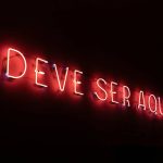 Neon sign at the entrance of a restaurant in São Paulo where it reads "deve ser aqui" ("it must be here")